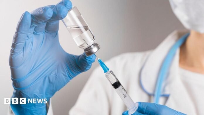 A doctor in a white coat and blue protective gloves holds a medicine in a syringe. The hand of the medical worker knocks on the syringe and knocks out the air with his finger.