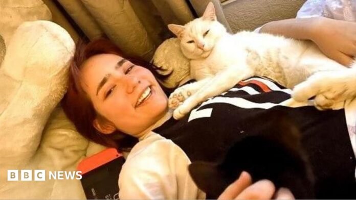 Elise Sebastian lies on a sofa with her cat on her. It is a white, short-haired cat. Elise is smiling and wearing a long-sleeved black and white top.