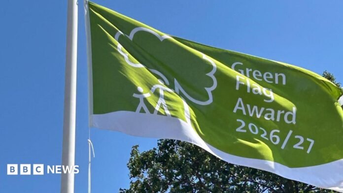 A green flag with a white stripe across the bottom and a tree with a parent and child stood next to it in white. It reads 'Green Flag Award 2026/27. A tree is in the background against the blue sky.