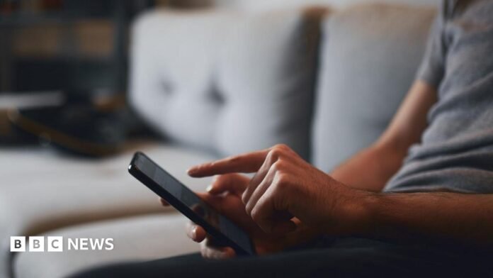 In this stock image, an unidentifiable man sits on a grey sofa, swiping on his smartphone.