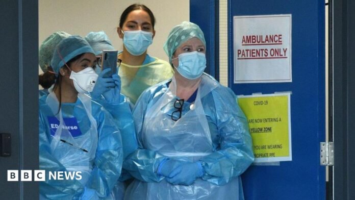 Four medics are standing at the doors of an emergency department in the pandemic. They are wearing plastic blue gowns and have face masks on. One has a sign on her chest saying "ED Nurse". 
