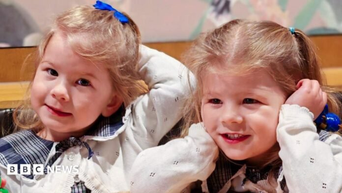 Nancy and Margo are photographed close up - their faces and blond hair in bunches are visible, and they are smiling. They are wearing the same clothes - a blue blouse and white cardigan.