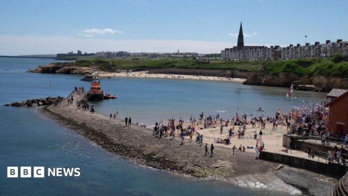 Lots of people on the sandy beach at Cullercoats on a sunny day. There are houses and a cathedral on top of the small cliff in the background.  A lifeboat is moored at the end of a stone jetty.