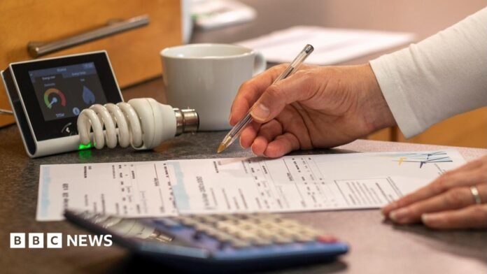 A person looks at their bills at home. A pen is in one hand and a bill lies on a table. There are other objects on the table, like a calculator, a meter, a bulb and a cup