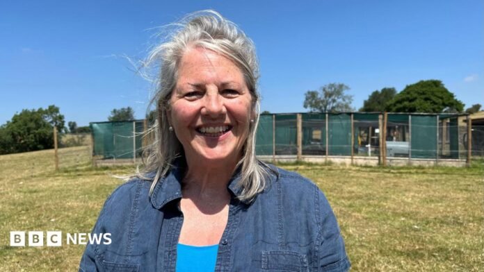 Perkins photographed looking into the camera and smiling. She is wearing a dark blue denim shirt with a bright blue vest underneath. She is stood on a large grassy field, with pens for the curlews behind her built out of chicken wire, wooden posts, and green material. The sky is clear and blue and there is strong sunshine. 