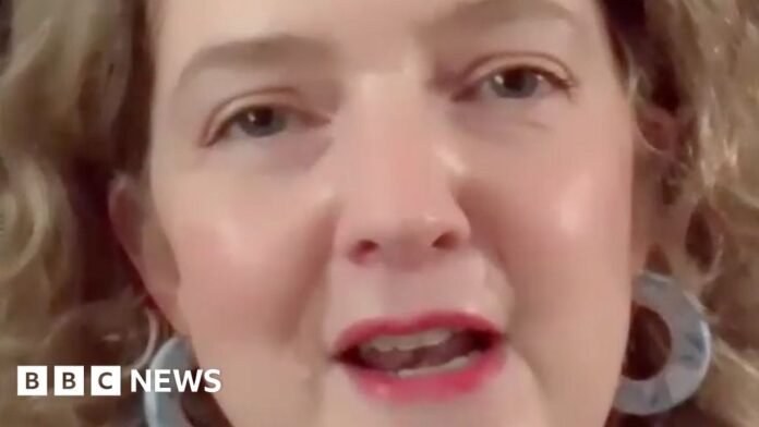 Frome and East Somerset MP Anna Sabine. She has shoulder length blonde-brown hair. She is wearing pink lipstick, a light green t-shirt and thick hooped earrings. There are books on a shelf behind her.
