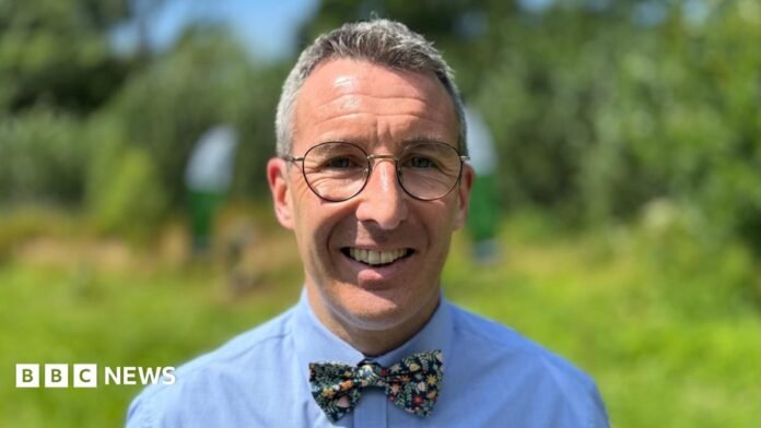 Andrew Muir has short grey hair and is wearing rounded glasses with a blue shirt and bow tie.