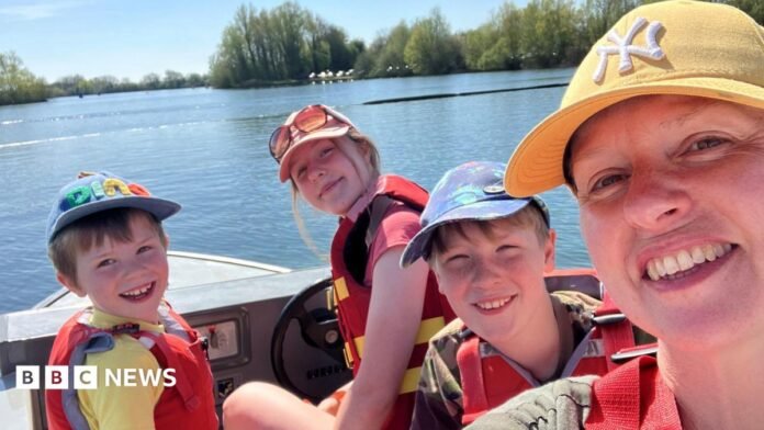 A woman wearing a yellow cap and three children, on a boat. All are wearing life jackets, and the photo is taken as a selfie-style with everyone looking at the camera