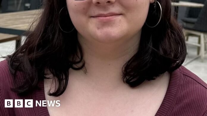 Aoísha is a young girl with shoulder-length dark hair. She is wearing a pair of wire glasses and large silver hoops. She is wearing a burgundy, ribbed shirt with buttons down the middle.