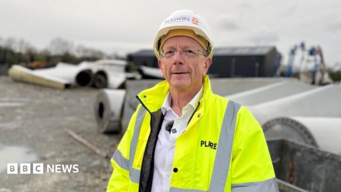 Andrew Billingsley, a man wearing frameless glasses, a white helmet, a yellow-high viz jacket and a white shirt, stands in an industrial yard looking at the camera.  There is a collection of large wind turbine blades behind him. 