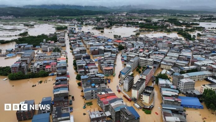 Aerial view of homes and buildings submerged in brown flood waters in Hengzhou, Nanning, China with a green mountain range and clody skies in the background