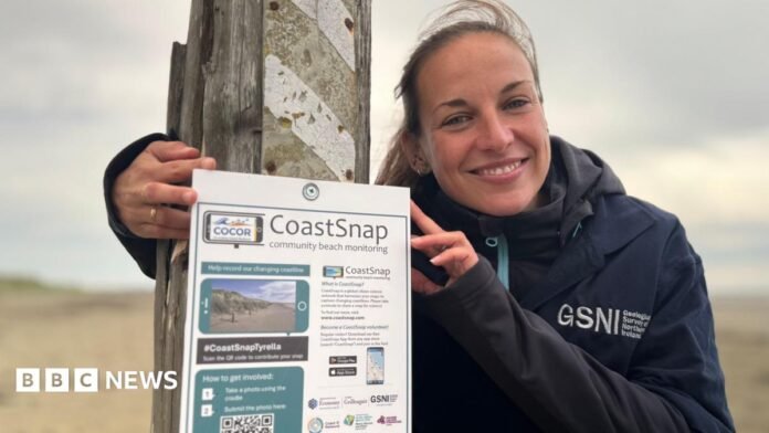 A lady with long brown hair tied back in a ponytail wearing a navy coat with GSNI written on it. She is standing beside a 'CoastSnap' poster which is pinned to a wooden post.
