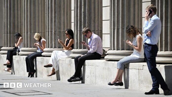 Five people sit along a low wall in the sunshine looking at their mobile phones,  while a man stands at the right-hand side speaking to someone on the phone