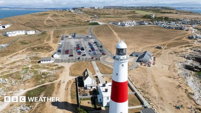 An aerial view shows Portland lighthouse next to a car park and a wide view of brown grass and vegetation, with the sea and a cloudy blue sky in the background, taken on Thursday.