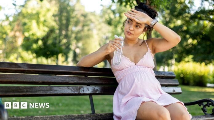 Pregnant woman sitting on a park bench in a light pink dress, holding a bottle of water.  