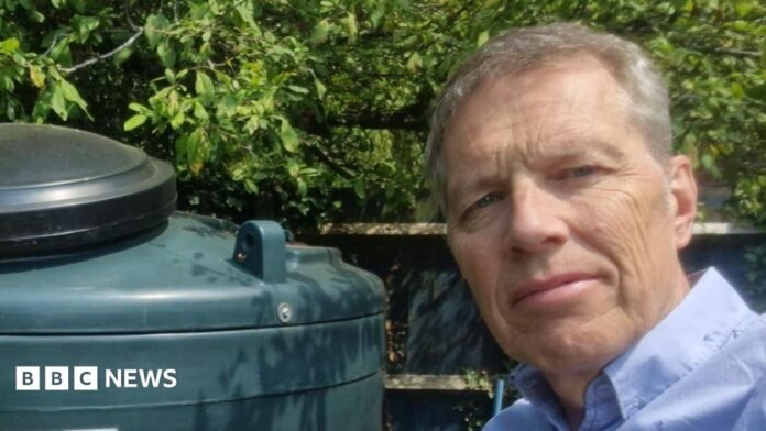A man in a blue shirt takes a selfie in a garden next to a green oil tank. He has grey hair and there are leaves behind the tank.