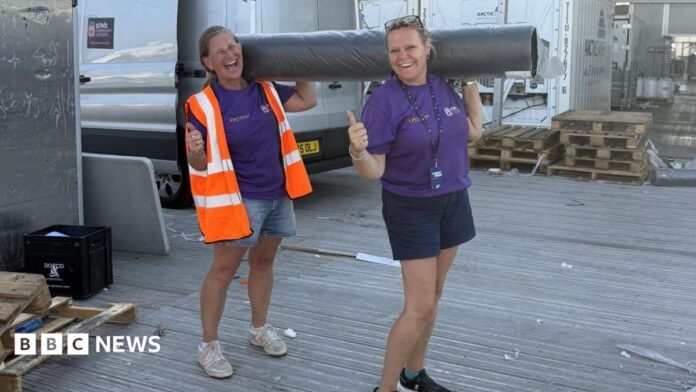 Two women, both holding their thumbs up, holding a carpet roll, both wearing shorts, and a purple T-shirt. The one on the left has on a high-vis jacket. A silver van is behind them and some wooden pallets. 