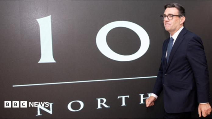Andy Burnham walks past a black painted wall bearing the words No 10 North in white writing. He is wearing a dark suit, white shirt and dark blue tie and black-framed glasses. He has a serious expression on his face.