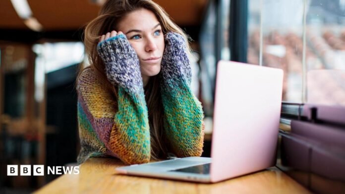 A bored young woman stares out of the window. She is sat in a library, using a laptop