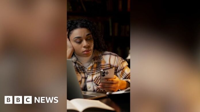 A woman checks her phone in the library. She is wearing a checkered shirt.