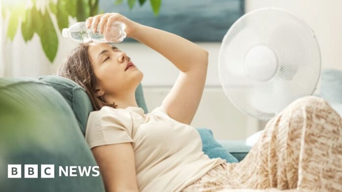 A woman reclines on a sofa next to a fan and with a bottle of water pressed to her forehead