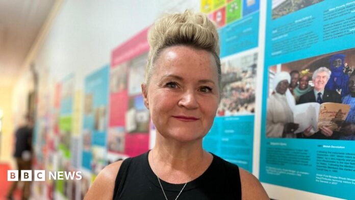 Donna is wearing a black sleeveless top and is smiling at the camera. She is stood in a corridor with images and articles in bright colours along the wall. She has short blonde hair which is shaved on the side and has a quiff on the top 