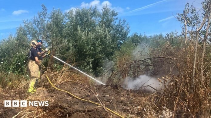 Photograph of a firefighter tackling a blaze at an illegal waste sump in Ashton-upon-Mersey. The image shows a jet of water being fired at the ground. 