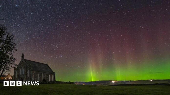 A night-time scene showing a church, which is darkness, and the sky behind the building glowing with a display of the aurora borealis. Stars shine behind the aurora's curtain if green and pink lights.