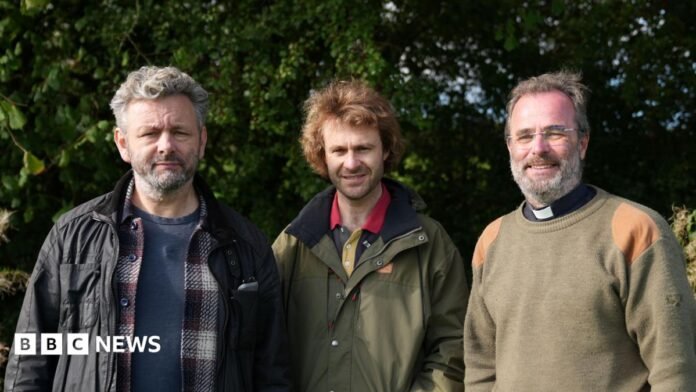 Three men sending side by side. First man has short grey hair and is wearing a blue t shirt, checked shirt and black jacket. The second man is wearing a khaki coloured jacket and the third man - a reverend - is wearing a khaki coloured jumper