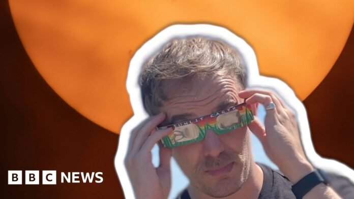 Man wearing a black tshirt and paper solar viewing glasses with a photo of a solar eclipse behind