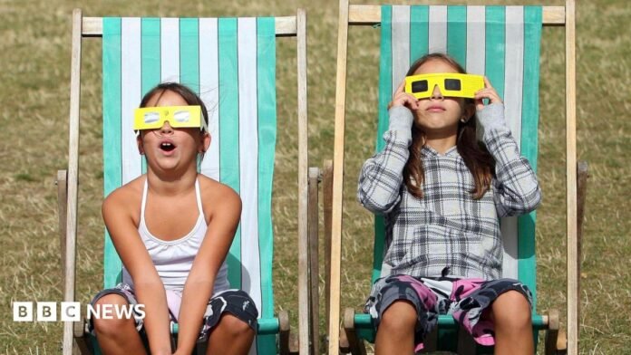 Two girls are sitting on green and white stripy deck chairs on grass wearing yellow eclipse glasses. They're looking up and it is a bright day.