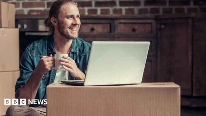 Smiling man sits on the floor surrounded by boxes, with a laptop on a box in front of him and a cup of tea in his hand.