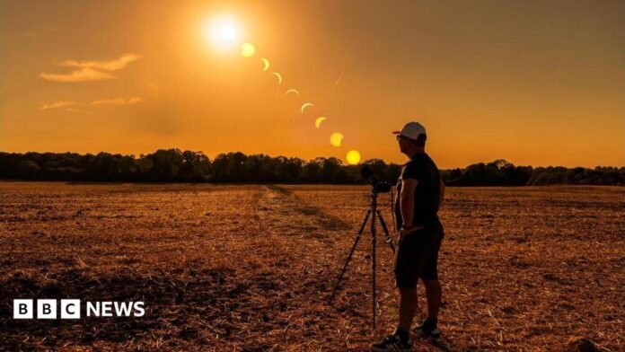 A person stands beside a camera on a tripod in a harvested field in Caterham, Surrey, during sunset. A sequence of the eclipse is shown across the sky, tracking the Sun as the Moon moves across it from high above the horizon to near sunset. Trees line the horizon beneath an orange sky.