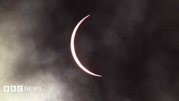 The moon has moved across the sun leaving a thin band of the sun visible which has taken on a pink hue. The sky is cloudy.