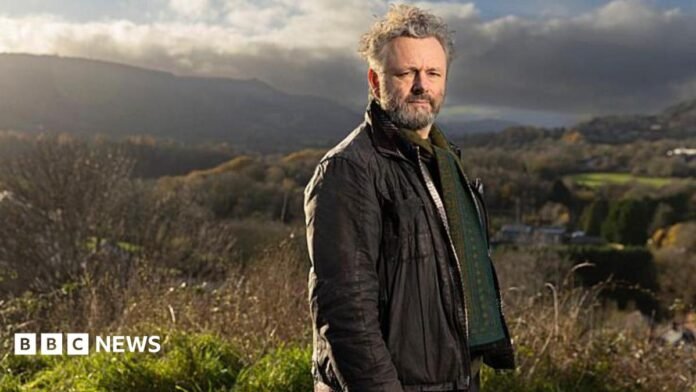Standing on high ground above a valley, Michael Sheen looks towards the camera with rolling hills and patchwork fields stretching into the distance. He is wearing a brown jacket with a green patterned jumper. Sheen has grey hair and facial hair. Strong afternoon sunlight and heavy clouds create dramatic contrasts across the rural landscape.