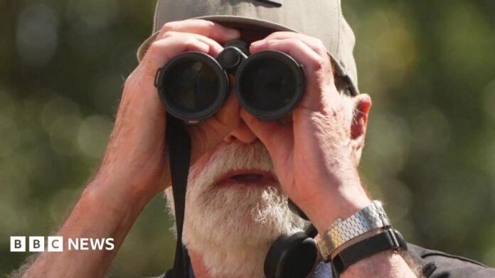 Nigel Hewitt is holding a black pair of  binoculars and wearing a cap. He has a white beard.