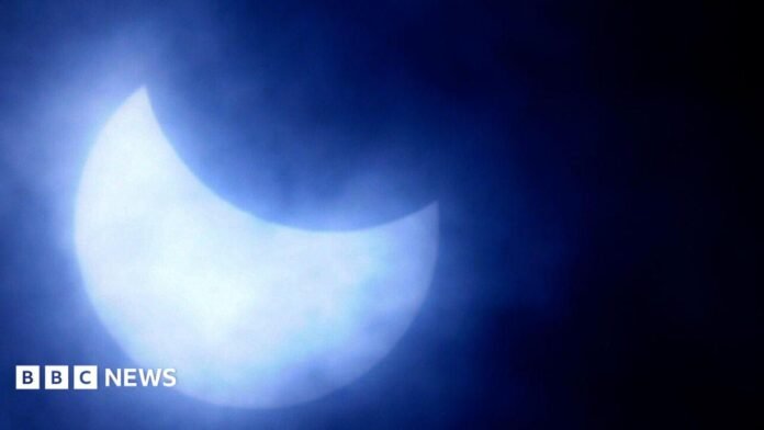 A view of the sun during a previous partial eclipse, which appears as a silvery-blue crescent against a black sky surrounded by a halo of light.