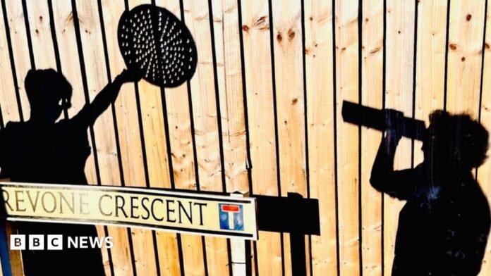 A wooden fence with two shadows cast upon it of people - one holding a cereal box to their eyes and peering through the opening, and the other holding a pizza tray with holes in above their head. There is a street sign at the bottom right of the image with the name 'Trevone Crescent' written on it.