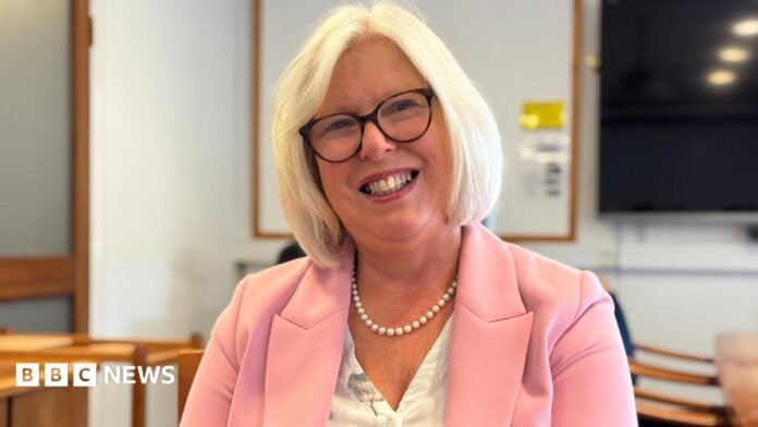Dr Nicola Brink is wearing a pink jacket as she sits in a room and smiling directly at the camera. She has pale blonde hair and glasses.