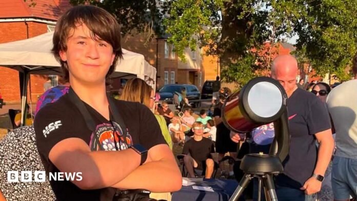 A young teenager is smiling and has his arms crossed stood next to a telescope