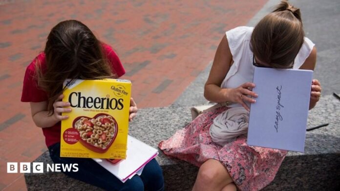 Two girls look into cardboard boxes, one a cereal box and another a shoe box, while sitting on a bench outside.