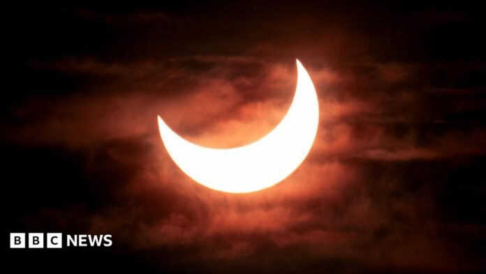 A partial solar eclipse showing the bright crescent of the sun as the moon moves across it from the top left corner. The sky is dark with clouds visible behind the sun
