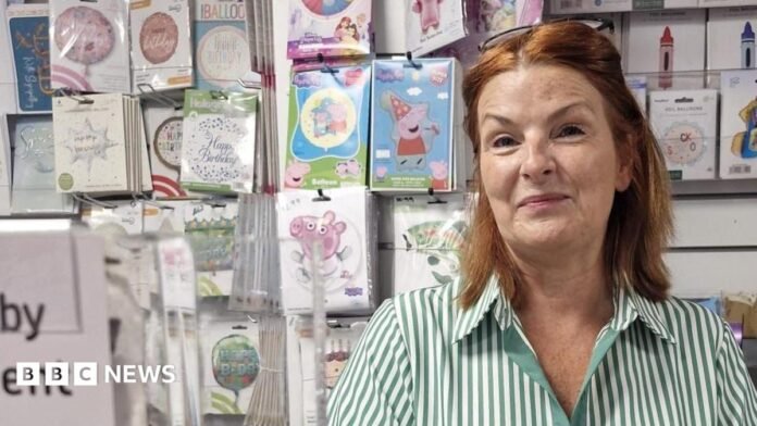 Karen Smith. She has shoulder-length copper-coloured hair and wears black glasses on top of her head and green and white striped blouse. She is standing behind the counter in her post office, with cards and decorations pictured behind her. She is smiling.