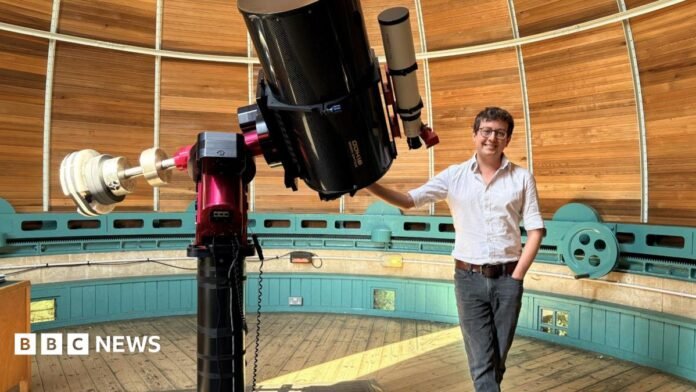 Dr Matt Bothwell standing next to a telescope. He is smiling and wearing a white shirt and black jeans. He is resting his hand on a large telescope. He is standing inside a circular wood-paneled observatory. 
