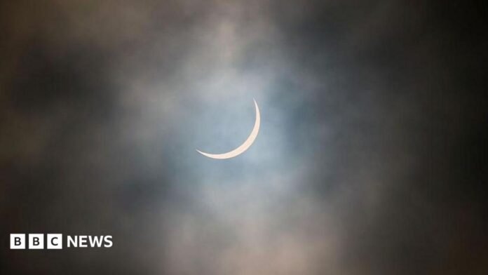 The sun is pictured during a partial solar eclipse. The sky is dark and cloudy. The sun is a thin arc in the sky.