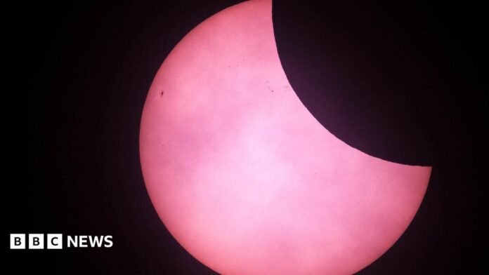 The top right corner of the sun has been obscured by the moon which appears black. The sun appears pink in the picture from St Martins in 2025.