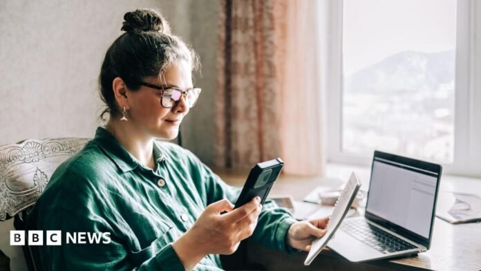 Woman sits at a window seat holding a phone and a pad with a computer on a table in front of her.